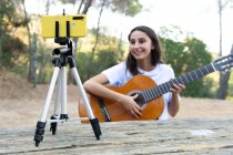 Весела жінка-підліток блогер грає на акустичній гітарі під час запису відео на мобільний телефон на тринозі в парку — стокове фото