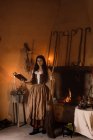 Ведьма в длинном платье смотрит в камеру с волшебной книгой заклинаний, стоя в уютной комнате с метлой и котлом — стоковое фото