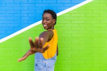 Vista posterior de la mujer afroamericana encantada de pie escuchar pared colorida y llegar a la mano a la cámara - foto de stock