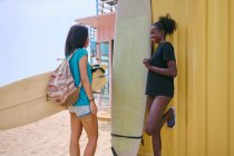 Zufriedene junge multirassische Sportlerinnen mit Longboard und Surfbrett stehen am Sandstrand vor einer Baustelle unter wolkenverhangenem Himmel und schauen einander an. — Stockfoto
