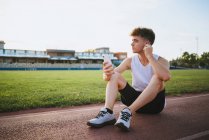 Contemplative young male athlete in undershirt with modern haircut listening to music from wireless earbud while looking away in stadium — Stock Photo