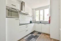 Interior de la elegante cocina con armarios blancos y alfombra de colores en parquet en el apartamento durante el día - foto de stock