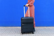 Seitenansicht einer nicht wiederzuerkennenden Frau im langen roten Kleid, die tagsüber mit Gepäck auf der Straße gegen eine blaue Wand steht — Stockfoto