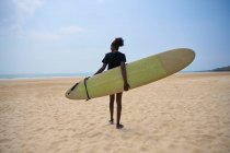 Visão traseira da atleta afro-americana com prancha de surf admirando o oceano da costa arenosa sob o céu azul nublado — Fotografia de Stock
