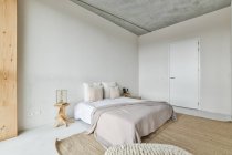 Креативный дизайн спальни с подушками и чехлом на кровати между дверью и столами на полу дома — стоковое фото