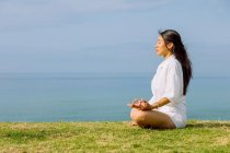 Vista laterale di sognante giovane femmina etnica con gli occhi chiusi meditando in posa Padmasana durante la pratica dello yoga sulla costa del mare — Foto stock