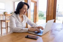 Happy mature female freelancer browsing on Internet on netbook working on new project while sitting at table at home — Stock Photo
