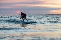 Surfista masculino de fato de mergulho e chapéu na prancha de remo surfando na praia durante o pôr do sol — Fotografia de Stock