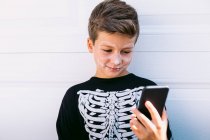 Веселый мальчик в костюме черного скелета с раскрашенным лицом на мобильном телефоне, сидя возле белой стены на улице — стоковое фото