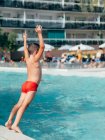 Full body back view of shirtless boy raising hands while push off edge of pool and jumping in water — Photo de stock