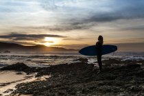 Seitenansicht einer unkenntlichen jungen Frau, die mit einem Surfbrett am Ufer steht, bevor sie während des Sonnenuntergangs am Strand in Asturien ins Meer steigt — Stockfoto