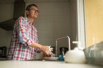 Вид збоку спокійного зрілого чоловіка, що миє брудні тарілки, стоячи біля раковини на кухні та роблячи домашнє завдання — стокове фото