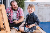 Hipster-Papa gießt Kräutertee aus Thermoskanne in Kalebassen-Kürbis gegen fröhlichen Jungen mit Bauholz auf Promenade — Stockfoto