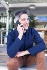 Glücklicher junger Mann in legerer Kleidung lächelt, während er auf der Bank sitzt und Telefonanrufe entgegennimmt — Stockfoto