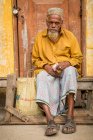 INDIA, BANGLADESH - DECEMBER 6, 2015: Senior ethnic elderly male in traditional clothes sitting on shabby weathered wooden door holding money and looking away — Stock Photo