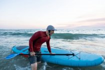 Side view of male surfer in wetsuit and hat carrying paddle board and entering water to surf on seashore — Stock Photo