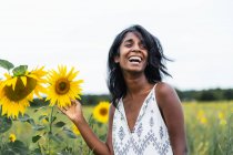 Sincero adulto fêmea étnica olhando para longe no prado tocando flores florescentes no campo no fundo borrado — Fotografia de Stock