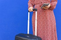 Cropped unrecognizable female in long red dress standing with luggage while tet message on smartphone on the street against blue wall in daytime — Stock Photo