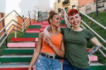 Модные счастливые гомосексуальные женщины, смотрящие друг на друга, спускаясь по лестнице к городским домам — стоковое фото