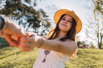 Sorridente adolescente donna in prendisole e cappello di paglia che tiene raccolto partner anonimo a mano mentre guarda la fotocamera nel parco — Foto stock