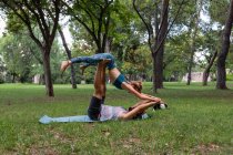 Vista lateral do casal flexível em sportswear praticando acroyoga juntos no tapete de ioga na grama contra árvores no parque durante o dia — Fotografia de Stock