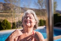 Mulher sênior alegre em biquíni desfrutando de salpicos de chuveiro perto da piscina com água clara transparente — Fotografia de Stock