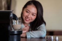 Colheita jovem etnia feminina apoiando-se em mãos contra cafeteira pod derramando bebida quente com espuma em vidro na cozinha da casa — Fotografia de Stock