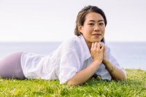 Искренне молодая этническая женщина в повседневной одежде смотрит в камеру, лежа днем на зеленом побережье — стоковое фото
