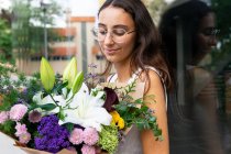 Encantadora joven hembra en gafas con flores en flor ramo contra la pared de vidrio en la ciudad a la luz del día - foto de stock