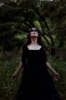 Mystische Hexe im langen schwarzen Kleid und mit aufgemaltem Gesicht, die in dunklen, düsteren Wäldern aufblickt — Stockfoto
