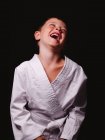 Lindo chico en kimono de karate riendo felizmente con la boca abierta en el estudio sobre fondo negro - foto de stock