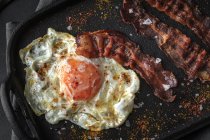Top view of sunny side up egg with fried bacon slices and condiments on tray against cutlery on dark background — Stock Photo