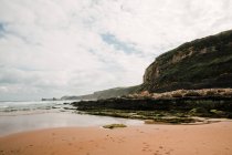 Vue spectaculaire de la montagne rugueuse avec mousse sur le rivage sablonneux contre l'océan sous un ciel nuageux en Cantabrie Espagne — Photo de stock