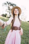 Sorridente adolescente donna in prendisole e cappello di paglia che tiene raccolto partner anonimo a mano mentre guarda la fotocamera nel parco — Foto stock