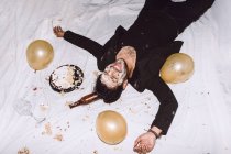 Drunk laughing male in smashed birthday cake lying near empty bottles from beer and balloons with eyes closed — Stock Photo