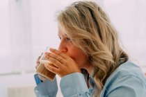 Вид сбоку взрослой женщины с вощеными волосами, пьющей вкусный кофе, глядя вперед в дом — стоковое фото