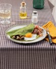 Hausgemachtes Frühstück aus Spinatpfannkuchen mit Speck, Ei und Pilzen, serviert auf einem weißen Teller mit einem Salz- und Ölstreuer auf einer karierten Tischdecke. — Stockfoto
