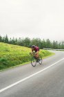 Спортсмен в защитном шлеме катается на велосипеде во время тренировки на асфальтовой дороге против зеленого холма и деревьев под легким небом — стоковое фото
