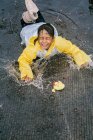 Affascinante bambino etnico in impermeabile che gioca con anatre di plastica che si riflettono nella pozzanghera increspata nel tempo piovoso — Foto stock