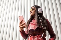 Baixo ângulo da moda jovem afro-americana de cabelos compridos em casaco vermelho e óculos de sol ouvindo música através de fones de ouvido e telefone celular enquanto desfruta de dia ensolarado na cidade — Fotografia de Stock
