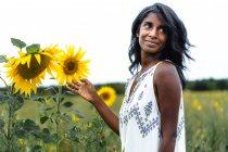 Aufrichtige erwachsene ethnische Frau, die auf der Wiese wegschaut und blühende Blumen auf dem Land auf verschwommenem Hintergrund berührt — Stockfoto