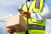 Cropped unrecognizable male worker in uniform carrying parcel while using mobile phone at work — Stock Photo