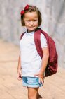 Vista lateral da criança em idade escolar com mochila no pavimento olhando para a câmera à luz do sol — Fotografia de Stock