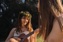 Adolescente en corona de flores jugando ukelele mientras se sienta contra la mejor amiga con auriculares inalámbricos en la espalda iluminada - foto de stock