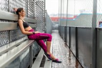 Visão lateral corpo inteiro de atleta feminina tonificada em activewear sentado no banco após o treino ao ar livre — Fotografia de Stock