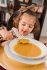Charmant enfant avec arc sur cheveux bruns et cuillère contre assiette de purée de courge soupe dans la maison — Photo de stock