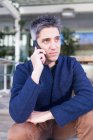 Serious young man in casual clothes while sitting on bench and answering phone call — Stock Photo