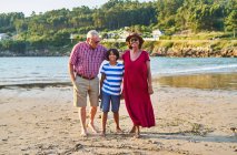 Full body of positive senior couple embracing boy while standing looking at camera on sandy coast of river in countryside — Stock Photo