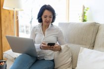Счастливая зрелая женщина фрилансер просматривает на смартфоне во время работы на ноутбуке сидя в диване дома — стоковое фото