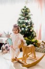Entzückendes kleines Mädchen, das neben einem hölzernen Schaukelpferd neben einem Weihnachtsbaum steht, der mit Lichtern und Spielzeug geschmückt ist — Stockfoto